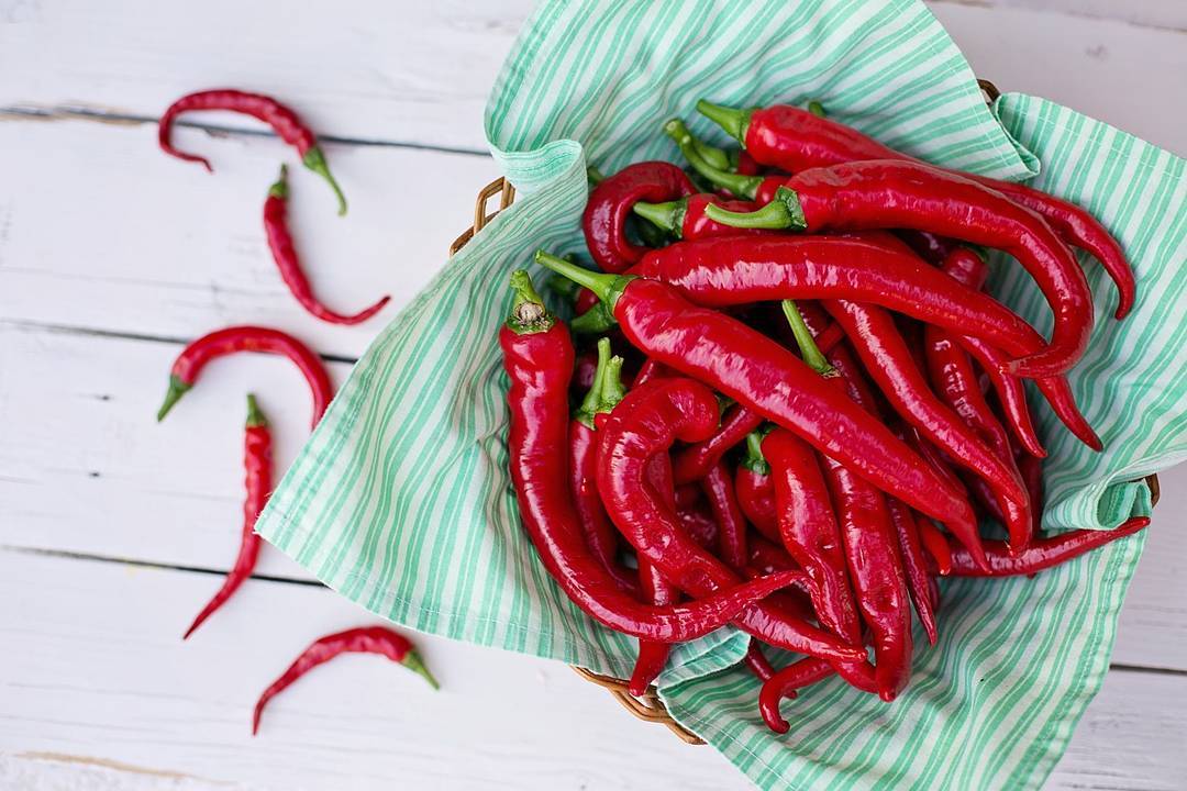 Basket of glossy red cayenne peppers on green striped cloth with additional peppers scattered on white wooden surface